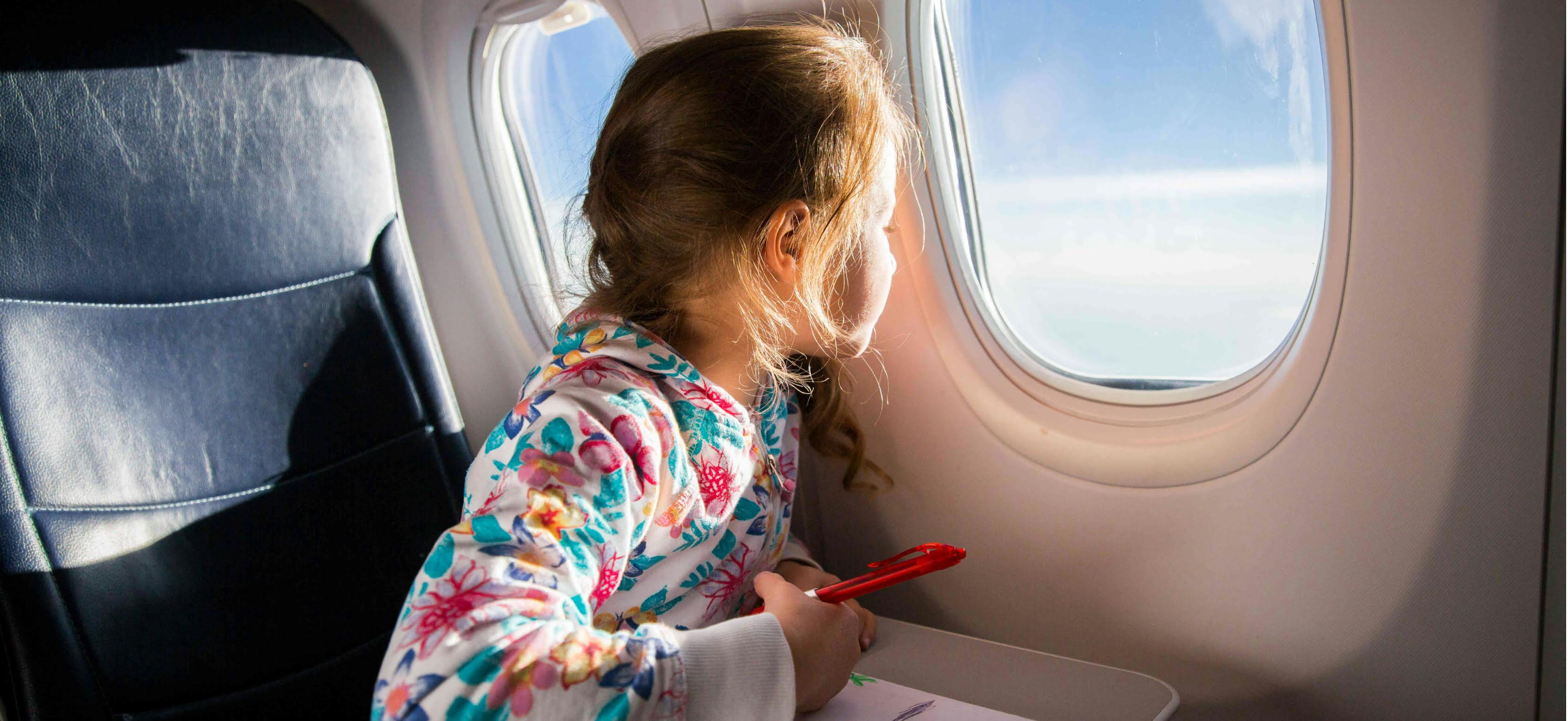 oung girl sitting on an airplane, gazing out the window while holding a red crayon and wearing a colorful floral hoodie.